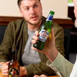 Three people seated at an indoor table. One man is holding a bottle of Peroni, whilst his two companions have their drinks on the table in front of them..  