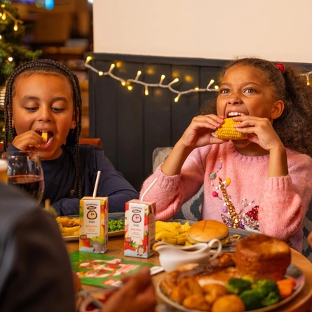 Two children sit at a festive dining table eating from plates of food, with one holding a piece of corn on the cob. The table is set with burgers, chips, vegetables, gravy, fruit juice cartons and drinks. Warm white fairy lights decorate the wall behind them, while a decorated Christmas tree with red and gold baubles and candy cane ornaments is partially visible to the side. Other diners are seated around the table in a cosy Christmas-themed restaurant setting.