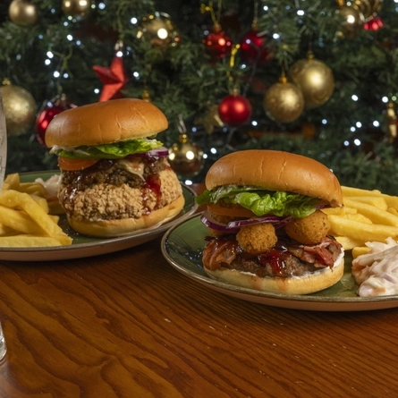 The Camembert Burger and the He's Bean Burger, each served on a plate with fries and coleslaw. The plates sit on a wooden table next to a glass of lemonade and a Christmas cracker, and a Christmas tree is visible in the background.