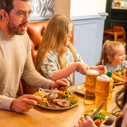 People sit at a wooden table in a pub dining area, eating a roast meal with meat, vegetables, potatoes, and Yorkshire puddings. Pints of lager and bottled juice drinks are on the table