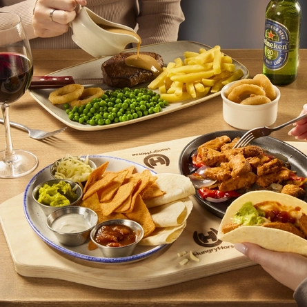 Two people sitting at a table, one of them is pouring sauce over a plate of steak, fries, peas, and onion rings, and the other person is using a fork to fill a fajita with food from the plates of meat, vegetables, nachos and sauce on the wooden board in front of them. A ramekin of onion rings, a glass of red wine and a bottle of Heineken 0.0% alcohol beer also sit on the table.