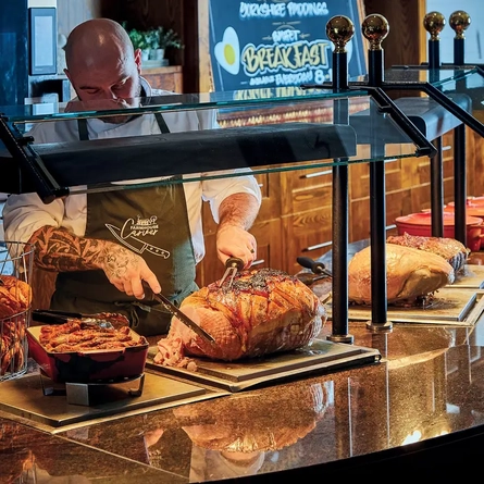 Carvery counter with food in dishes under a hot plate. Meat being carved by staff