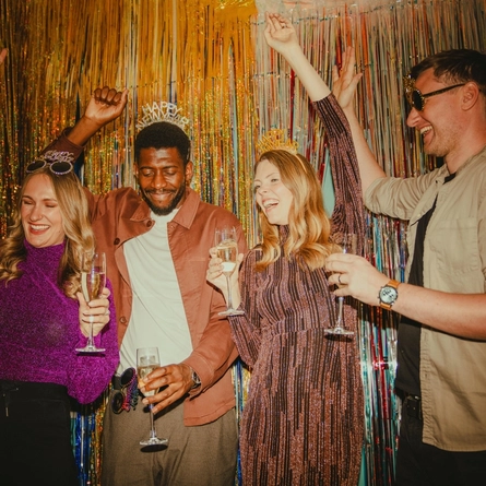 Four adults stand in front of a sparkling gold and silver tinsel backdrop, raising champagne flutes in a New Year’s Eve toast. Several people wear “Happy New Year” headbands, while one person wears novelty sunglasses on their head. The festive party scene features colourful metallic decorations, evening attire and celebratory drinks.