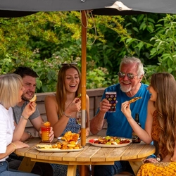 A mixed group of people seated around a wooden table, under an umbrella in a beer garden, enjoying a variety of sharers and drinks.