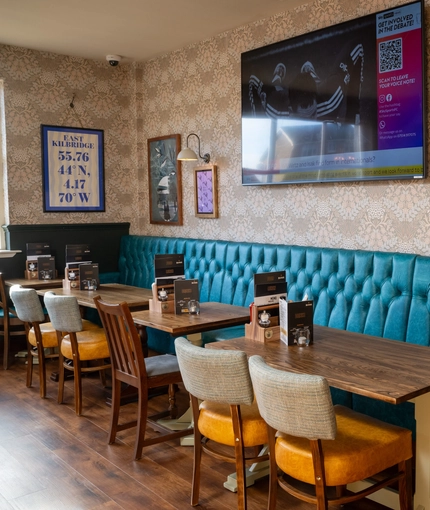 The interior restaurant seating area at The Kittoch with a long, upholstered booth seat, a TV on the wall above it, and framed artwork on the walls.