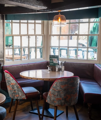 An interior restaurant seating area at The Ship; with wooden beams on the walls and ceiling, and padded bench seating in the large bay window.