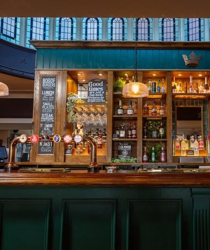 A close up view of the wood panelled bar inside the Golden Hind, with glasses, bottles of wine, and bottles of alcoholic spirits on wooden shelves behind the counter. Leaded windows are visible through an oval opening in the ceiling above the bar.