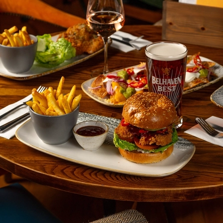 A round, wooden restaurant table laden with plates of food including burgers, fries, nachos, and salad. A glass of wine, a glass of Belhaven Best ale, a glass of Belhaven Twisted Thistle IPA, cutlery, and menus also sit on the table.