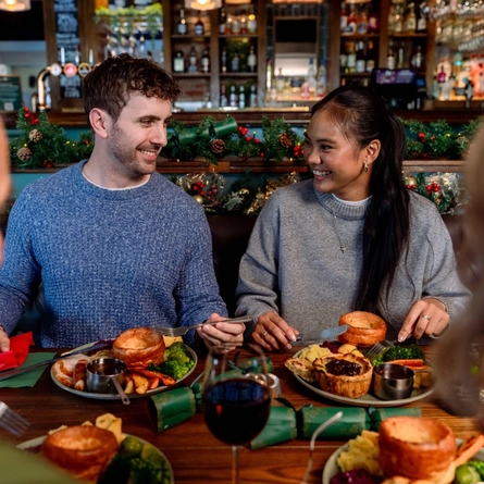 An image focusing 2 people sat within a group at a table within the interior restaurant seating area enjoying drinks and dishes from the Christmas menu at a Community Social venue.