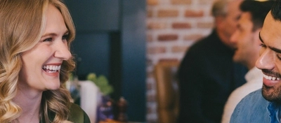 A man and woman seated at an indoor wooden table with plated food in front of them.  They are both holding drinks in a cheers motion.
