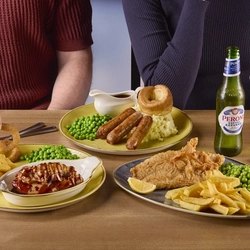 A plate of fish, chips, peas, and lemon, another plate of sausages, mashed potatoes, peas, Yorkshire pudding, and a jug of gravy, and another plate of chicken in a small roasting dish, fries, onion rings, and peas. The three plates sit on a wooden table along with cutlery, a glass of fizzy drink, and a bottle of Peroni lager.