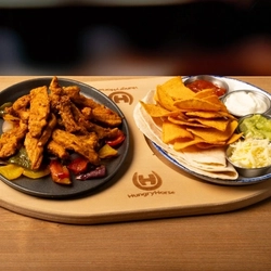 A wooden board holding a skillet with chicken and vegetables and a plate with a flatbread, nachos and dips on a wooden table.