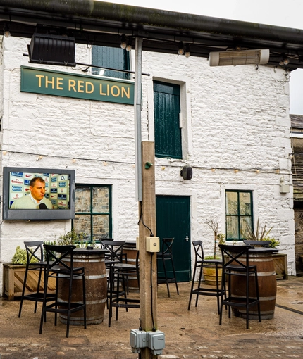 An exterior seating area at The Red Lion, with wooden tables, a TV on the wall, and string lights.
