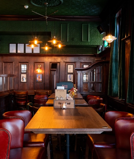 The bar and interior restaurant seating area at the King's Head, with dark wood panelled walls; green velvet curtains; upholstered chairs; and green, textured wallpaper on the ceiling.
