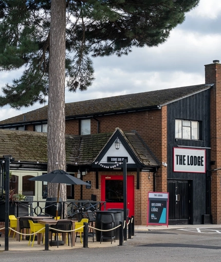 The exterior facade, signage, and beer garden seating area of The Lodge in Alvaston, with a large tree in front of the building, shade umbrellas on some of the tables, and accessible parking near the entrance.