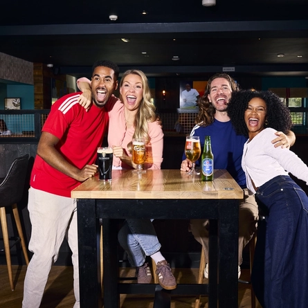 An image showing 4 friends at a table enjoying drinks and celebrating while watching on a football match within the interior seating area at a Flaming Grill venue.