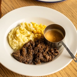 Haggis, Neeps and Tatties served on a plate with a small pot of sauce. The plate sits on a wooden table next to a glass of Belhaven Best ale and another plate of food.