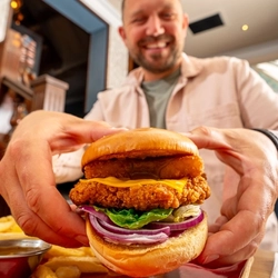 A close up image of a burger with a person in the background within the interior restaurant and seating area at a Hungry Horse venue.