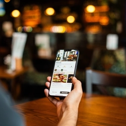 An over-the-shoulder view of a person at a wooden table holding a smartphone in one hand. The phone screen shows a venue page on the Greene King app.