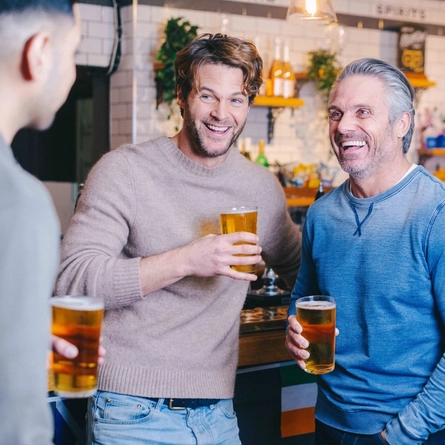 Three people standing at the bar inside a pub, each holding a glass of cider.