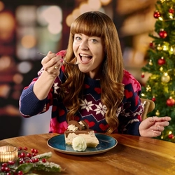 A close up view of a person sitting at a restaurant table and holding up a spoonful of the cheesecake served on the plate in front of them. A tealight surrounded by berries and pine cones sits on the table; and a Christmas tree is visible in the background.