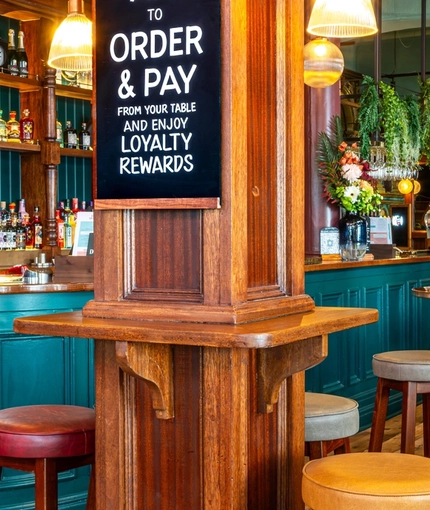 The bar and interior restaurant seating area of The Fountains Abbey, with wooden and metal tables, upholstered seating and a TV on the wall.