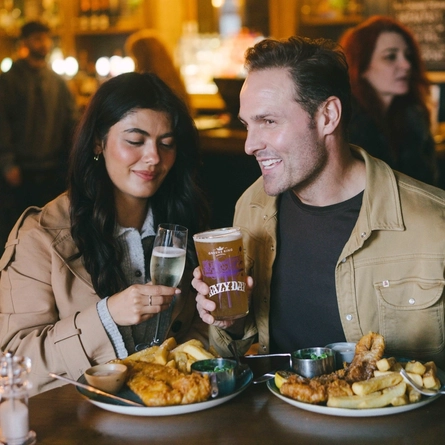 An image of a 2 friends enjoy Fish & Chip dishes with drinks sat within the interior restaurant seating area at an Urban Core Venue.