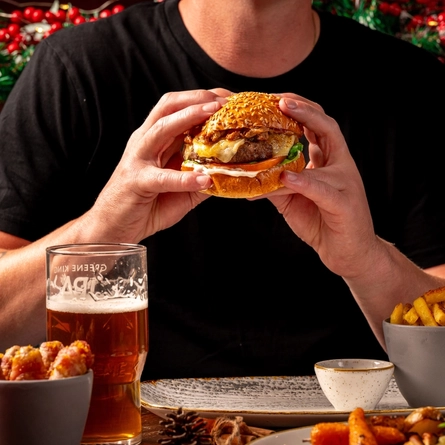 A man is seated at a wooden table with festive decor, he is holding a burger in his hands.  Only the torso and arms are visible in the picture.