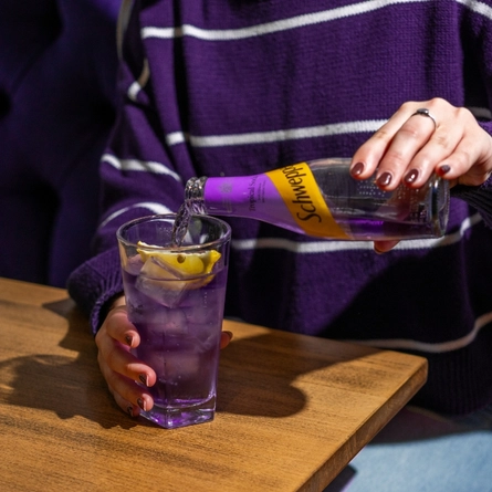 A woman seated at an indoor wooden table, pouring from a bottle of Schweppes Tropical Soda into a highball glass.  Only the hands and torso are shown in the image.