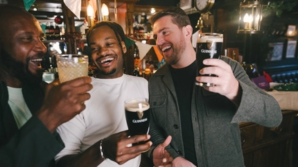 Three people laughing together inside a pub; one of them is holding a cocktail, the other two are holding glasses of Guinness.