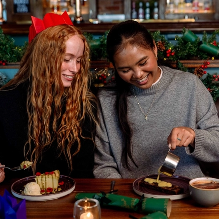 An image focusing 2 people sat within a group at a table within the interior restaurant seating area enjoying drinks and dishes from the Christmas menu at a Community Social venue.