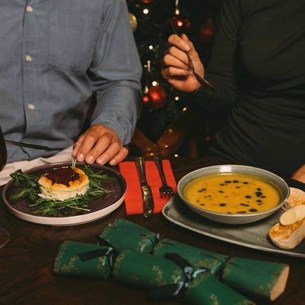 A bowl of soup served on a plate with two slices of bread topped with butter, sitting on a wooden restaurant table along with another plate of food. Cutlery, serviettes, a glass of white wine, a cocktail, and two green Christmas crackers also sit on the table; and two people are sitting at the table, one holding a knife, and the other holding a spoon.