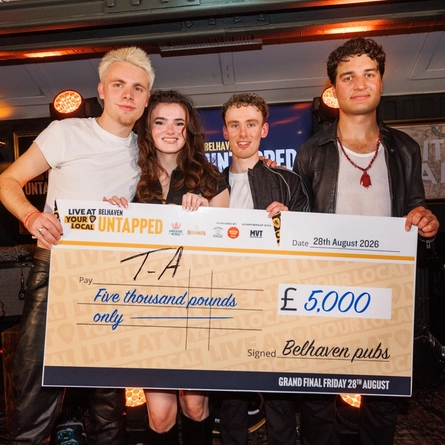 The four members of T-A who are the first ever winners of the Belhaven Untapped competition. The band is standing on a stage at The City Walls in Stirling, holding a giant cheque for £5000.