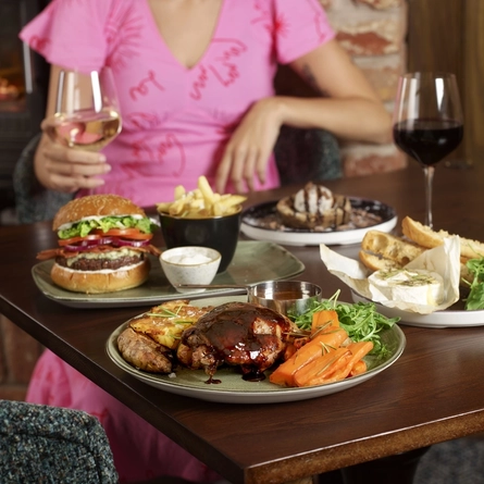 A person in a pink dress holds a glass of white wine at a table with assorted dishes including a burger, grilled meat, bread with cheese, and dessert.
