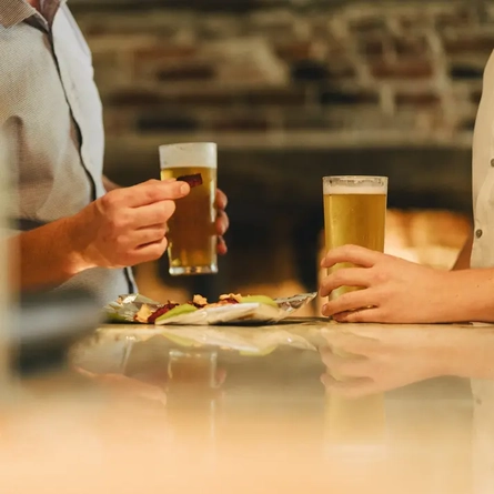 Two pints of beer, one being held and one sitting on a table with a hand around it, and an open bag of crisps between them.