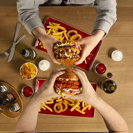 The hands of two people seated at a wooden table each holding a burger.  On teh table in front of each person are plates of fries and onion rings.