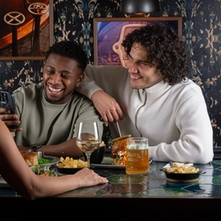 Three people sit around a restaurant table. One person leans toward another while showing something on a phone. The person beside them has an arm around their shoulders. Across the table, another person sits facing them. The table is filled with dishes including fries, salad, and a main dish, along with drinks such as a glass of white wine and a pint of beer.