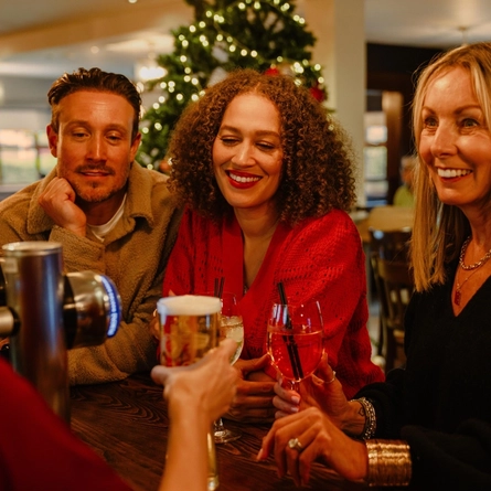 Three people sit together at a bar in a festively decorated pub, holding drinks and looking towards someone off camera. Glasses of rosé wine, a pint of beer and a mixed drink are arranged on the wooden bar, while a Christmas tree with warm fairy lights glows in the background, creating a cosy seasonal atmosphere.
