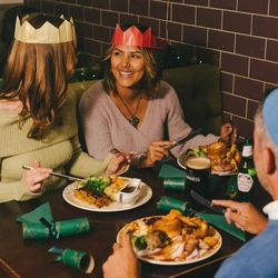An image of a group of friends sat at a table enjoying festive main dishes and various drinks within the interior restaurant seating area at a Proper Local venue.