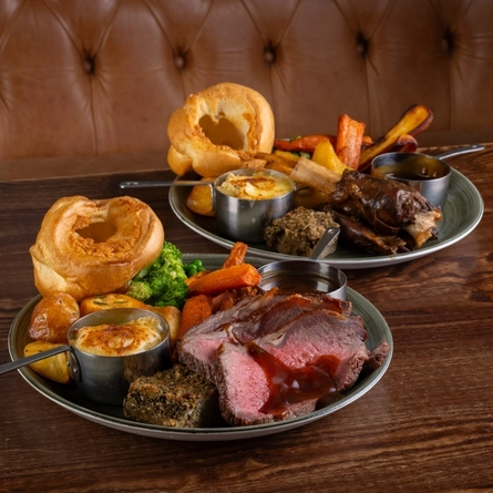 Two plates of roast dinner, including meat, Yorkshire pudding, vegetables and gravy, sitting on a wooden table.