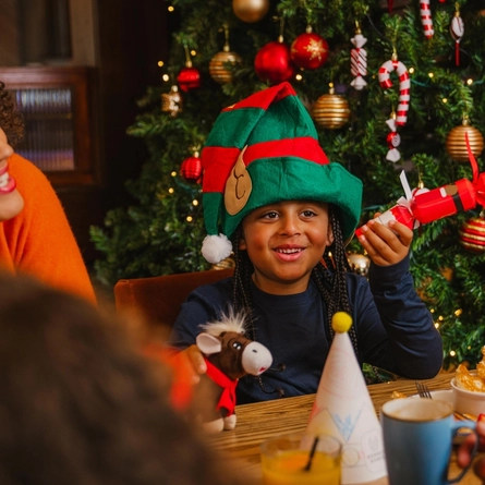 A child wearing a green-and-red elf hat sits at a wooden table in front of a decorated Christmas tree, holding a red Christmas cracker. The table is set with breakfast dishes, a glass of orange juice, a juice carton, a paper party hat and a small reindeer toy. Two other people are partially visible at the table, while the tree behind is decorated with red and gold baubles, candy cane ornaments and warm fairy lights.