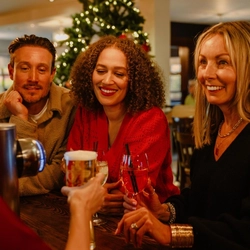 Three people sit together at a bar in a festively decorated pub, holding drinks and looking towards someone off camera. Glasses of rosé wine, a pint of beer and a mixed drink are arranged on the wooden bar, while a Christmas tree with warm fairy lights glows in the background, creating a cosy seasonal atmosphere.