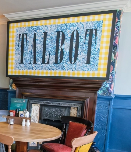 An interior restaurant seating area of The Talbot Inn, with framed artwork and potted plants on the walls, and a fireplace.
