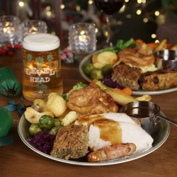 A close up view of a Roast Turkey dinner, featuring slices of roast turkey, a Yorkshire pudding, roast potatoes, vegetables, pigs in blankets, and gravy, served on a plate. The plate sits on a table with a glass of Level Head IPA, another plate of food, and Christmas table decorations. 
